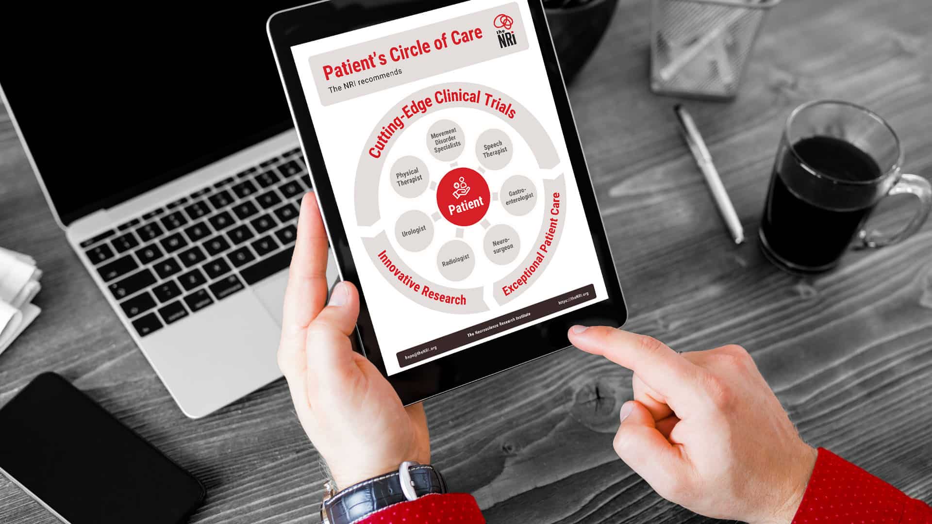 Foreground: hands holding an iPad with The NRI's Circle of Care document on the screen. In the background is a desk featuring a laptop, pen, phone, and cup of coffee.