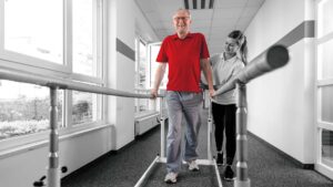 Older male patient walking with the assistance of parallel bars and a physical therapist